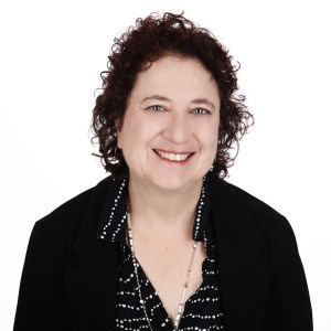 Headshot of Elana Varon, smiling. She is wearing a black blazer over a navy blue shirt with white stripes and a gold necklace with white beads